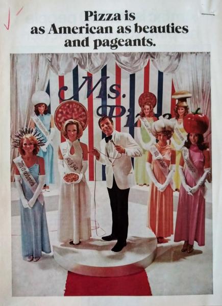 A master of ceremonies is surrounded by beauty pageant contestants with pizza ingredient hats.